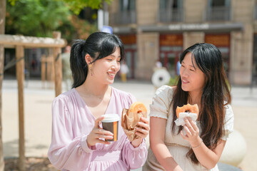 Two women are sitting on a bench and eating donuts. One of them is holding a coffee cup