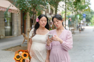 Two women are walking down a street, one of them looking at her phone. The other woman is holding a bouquet of flowers