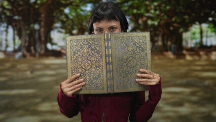 Young hispanic woman holds an ornate gold embossed leatherbound book with hands amidst dense leafy forest under soft natural light; curiosity.