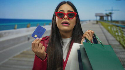Woman shopping seaside promenade holding bags and credit card looks worried while enjoying outdoor...
