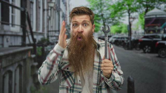Man holding wrench with one hand and raising other hand on street, wearing plaid shirt and sporting a long beard; surprise. - Powered by Adobe