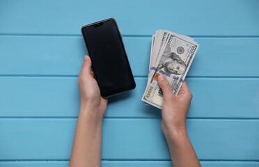 Woman with money and smartphone at blue wooden table, top view