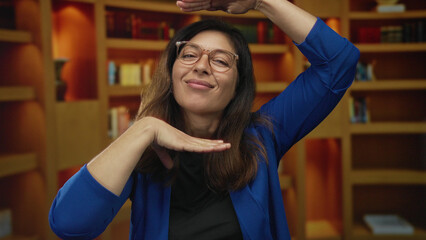 Woman in blue cardigan frames face with hands and smiles in front of library bookshelves; creative vision joy.