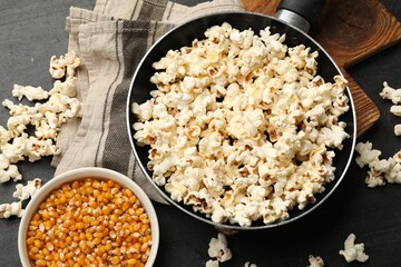Tasty popcorn in frying pan and corn kernels on black table, flat lay