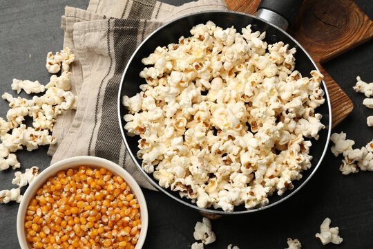 Tasty popcorn in frying pan and corn kernels on black table, flat lay