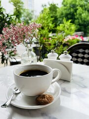 A white cup of black coffee with a cookie on a saucer stands on a marble table in a cozy outdoor café surrounded by greenery. Summer atmosphere with flowers and blurred city background.