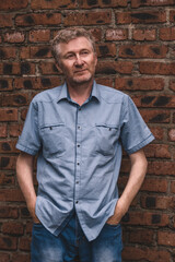 authentic portrait middle-aged man standing casually against old brick wall, hands in his pockets, wearing blue checkered shirt. he appears pensive, reflecting urban life around him. close up.