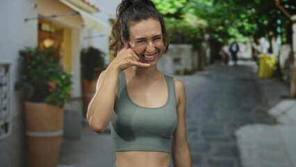 Young hispanic woman smiling and making call sign with her hand in sunlit bustling urban street; playfulness.