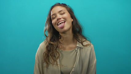 Obraz premium Woman sticks out tongue with long hair loose over blue studio wall, wearing beige blouse; playfulness.