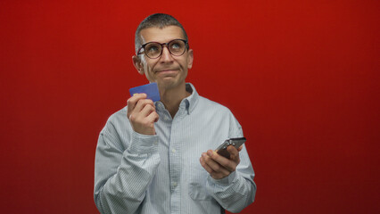 Man holding credit card and smartphone against a vivid red background, suggesting a financial decision or online shopping scenario with focus on technology and finance.