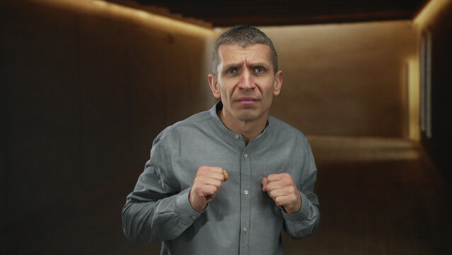 Man in grey shirt standing in dimly lit indoor room with serious expression and clenched fists, conveying tension or assertive mood.