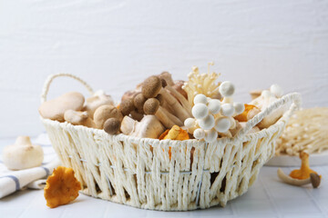 Different raw mushrooms in wicker basket on white tiled table, closeup