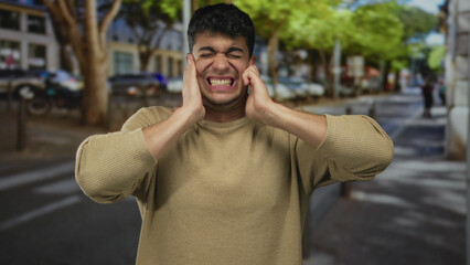 Man shields ears covering street commotion, guy young hispanic urban hands raised against outdoor...