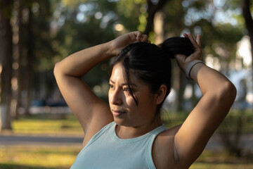 Latina athlete gathering long hair into ponytail, preparing for outdoor fitness routine amid sunlit...
