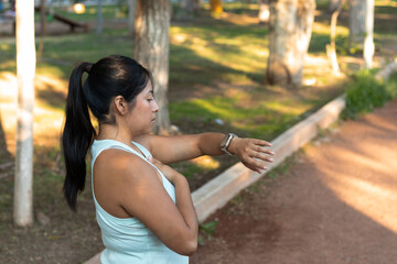 Latin fitness enthusiast stretching arm, reviewing smartwatch data during morning workout in sunlit park, embodying wellness and summer fitness goals