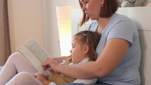 Young woman shares a quiet reading moment with her daughter in bed. Educational bonding and speech development during relaxed indoor time. Mid shot