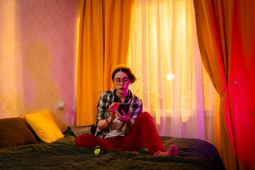 Relaxed teenage girl using phone in cozy bedroom with orange curtains. Concept of youth leisure and mobile tech. Mid shot