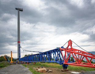 A tall wind turbine tower under construction with large cranes and steel framework on a cloudy day at an industrial worksite.