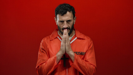 Hispanic man in orange prison jumpsuit standing against a red background with hands in prayer.