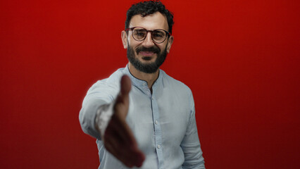 Man with beard and glasses extending hand for handshake against vibrant red background symbolizing greeting and welcome in a positive professional context.