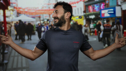 Fototapeta premium Hispanic man with beard gestures playfully on a busy street with colorful flags, surrounded by blurred crowd in an urban outdoor scene during daytime.