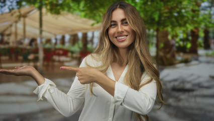 Fototapeta premium Blonde woman with outstretched hand and finger pointing to palm at outdoor cafe patio; invitation welcome hospitality offer.