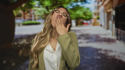 Fototapeta premium Woman with glasses in business suit yawns covering mouth on a downtown street; morning fatigue routine.