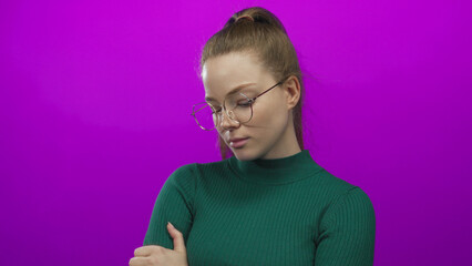 Young woman wearing green turtleneck and glasses crosses arms and looks down in studio with purple wall; rejection.
