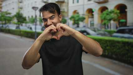 Fototapeta premium Young man making heart shape with hands on city street, smiling brightly, surrounded by urban buildings and greenery, depicting a joyful outdoor moment.