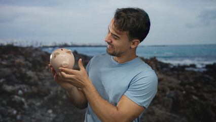 Young hispanic man smiling, holding piggybank at seaside with ocean view in background showcasing savings and leisure at beach.
