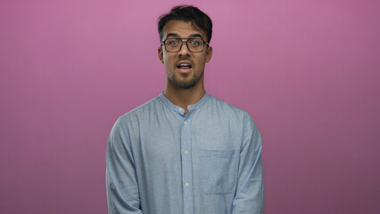 Obraz premium Young hispanic man wearing glasses and blue shirt, expresses surprise and excitement over pink background using facial expressions and gestures.