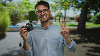 Young hispanic man smiling at camera in a park holding a camera with a peace sign, surrounded by vibrant green nature during daylight.