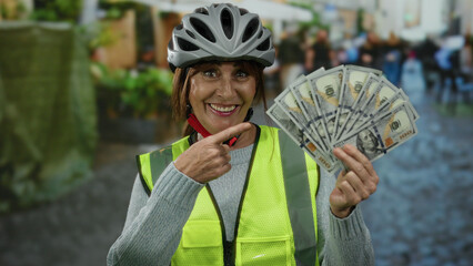 Woman cyclist in city wearing helmet and reflective vest holding and pointing at american dollar...