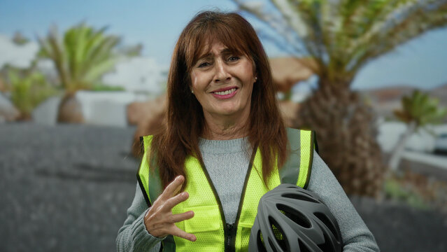 Senior hispanic woman standing outdoors on a city street wearing a safety vest and holding a helmet, with palm trees in the background.