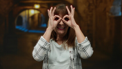 Senior hispanic woman playfully mimicking binoculars with hands on a dimly lit city street,...
