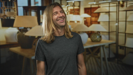 Blond man smiling beside lamp display on wooden table inside a building with shelves in view;...