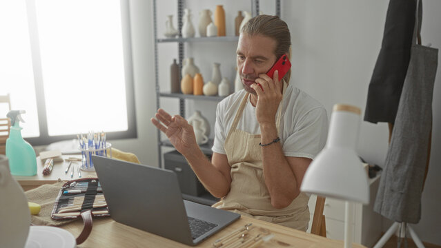 Man holds smartphone to ear while sitting at pottery studio desk with laptop and brushes; concentration. - Powered by Adobe