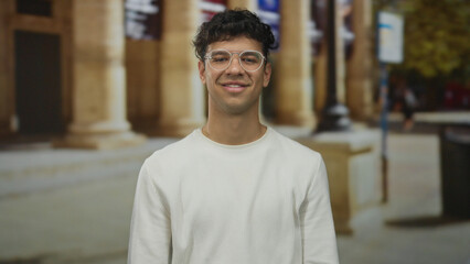 Man with arms crossed wearing glasses and smiling while winking on street by building columns; confidence.