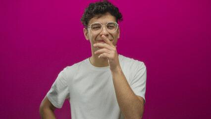 Young man with glasses wearing white t shirt facing camera and hand to mouth laughing in magenta...