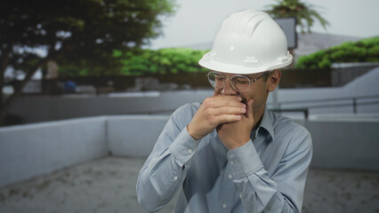 Man wearing hardhat covers mouth with hands in a cough gesture beside a basketball hoop on an outdoor building court; safety.