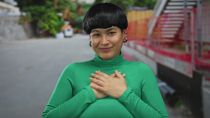 Woman outdoors wearing green sweater with hands on chest smiling on a quiet urban street, surrounded by greenery and urban structures, exuding warmth and positivity.