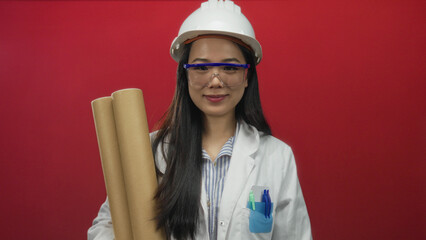 Young chinese woman architect in hardhat holds blueprints against red background, wearing safety goggles and white coat.