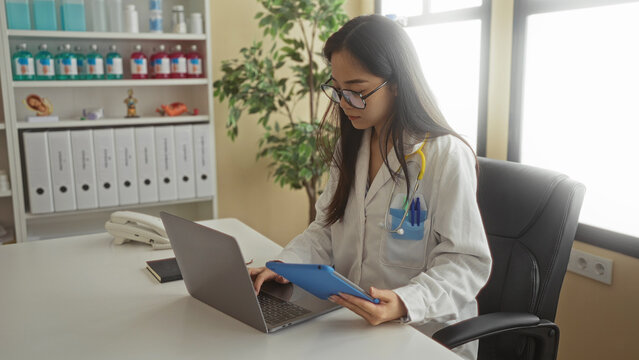 Young chinese woman doctor in a clinic room, wearing a stethoscope and uniform, works on a laptop and tablet, surrounded by medical supplies and documents. - Powered by Adobe