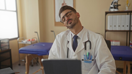 Man wearing white coat and stethoscope pauses typing with head tilt and eyes closed in a building;...