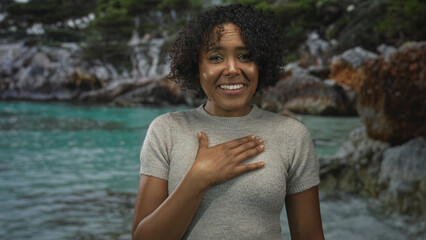 Woman smiles and holds hand on chest in natural forest setting while looking at camera with relaxed...