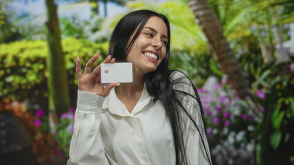 Fototapeta premium Woman smiling holding card in outdoor park setting with tropical plants and flowers, showcasing a cheerful and relaxed atmosphere.