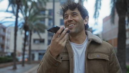 Hispanic man speaking on smartphone outdoors on a city street lined with palm trees, exuding...