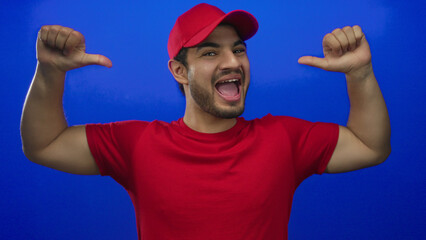 Naklejka premium Hispanic courier man wearing red shirt and cap points thumbs in studio with open mouth expression and bright blue backdrop; pride.