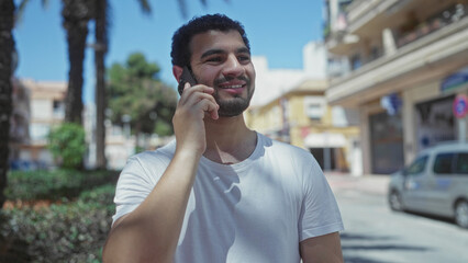 Man in white t-shirt holding smartphone to ear on a sunny urban street lined with palm trees; connection.