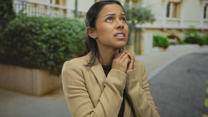 Hispanic woman in a beige jacket appears anxious standing on a quiet street, outdoors, her expression conveys fear and worry, surrounded by greenery and elegant architecture.